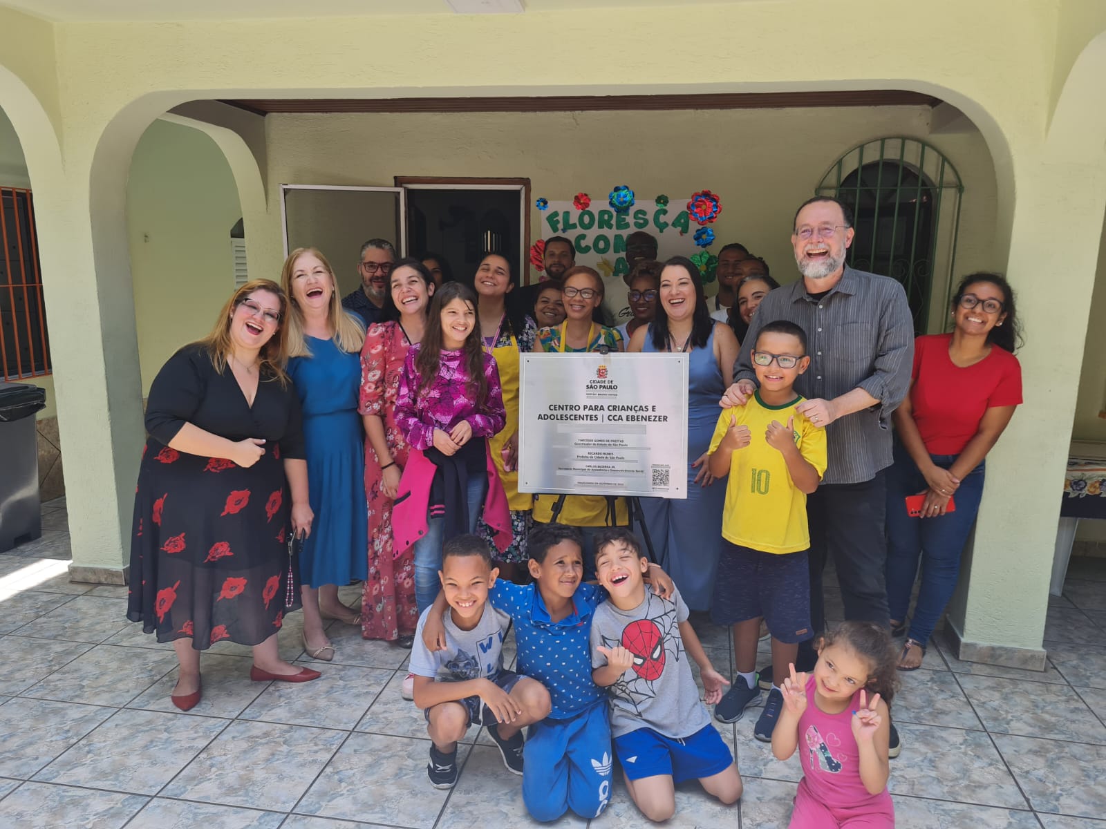 Diversas pessoas sorriem e posam para a foto no meio de uma sala de paredes amarelas. No meio, há uma placa, na qual se lê: Centro para Crianças e Adolescentes | CCA Ebenezer.