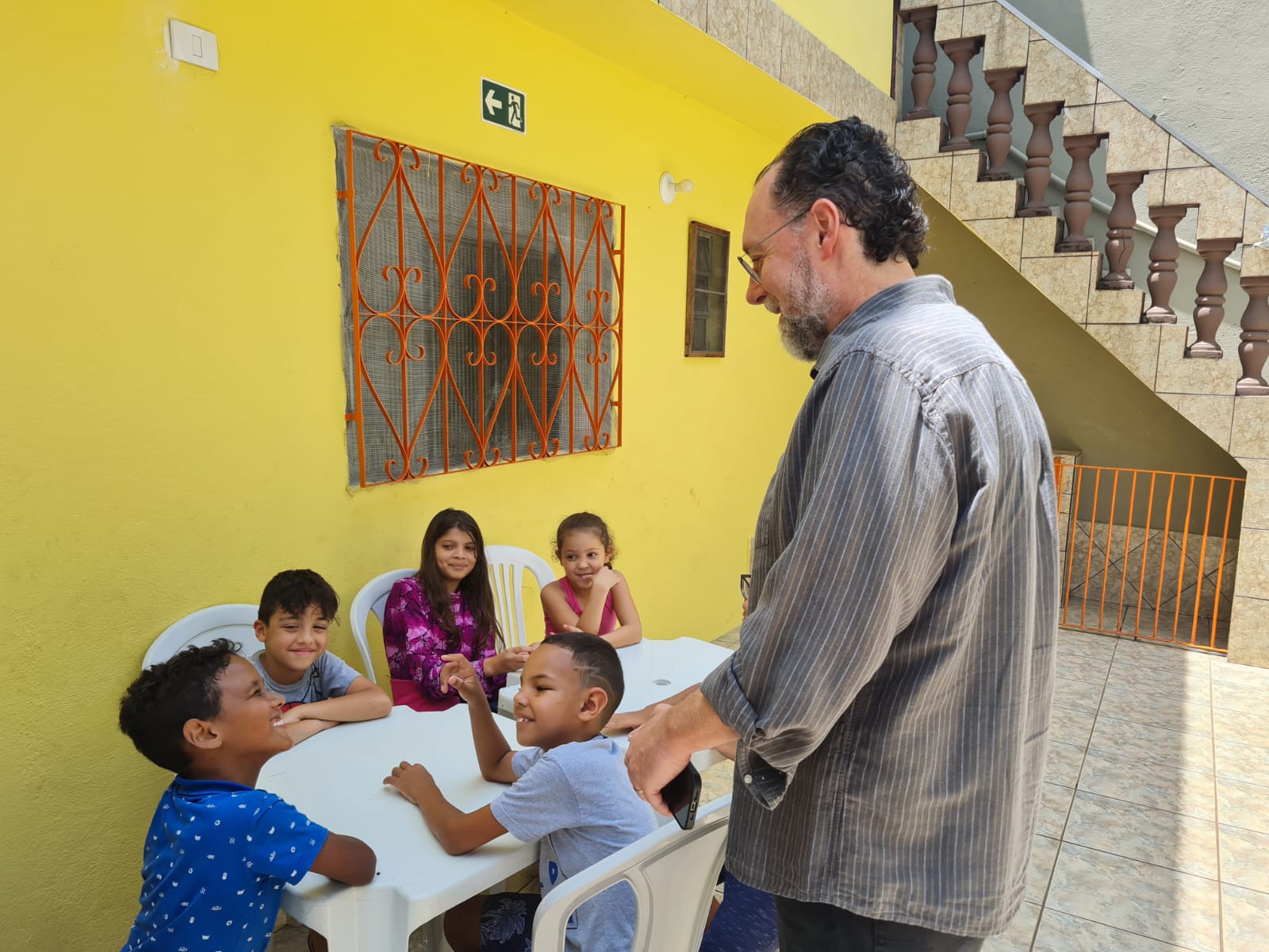 Cinco crianças se sentam em cadeiras de plástico brancas, em frente a uma mesa de plástico branca. Atrás delas, a parede é amarela e tem uma janela de vidro com grades laranjas. Em pé, na frente, há um homem branco de camisa cinza.