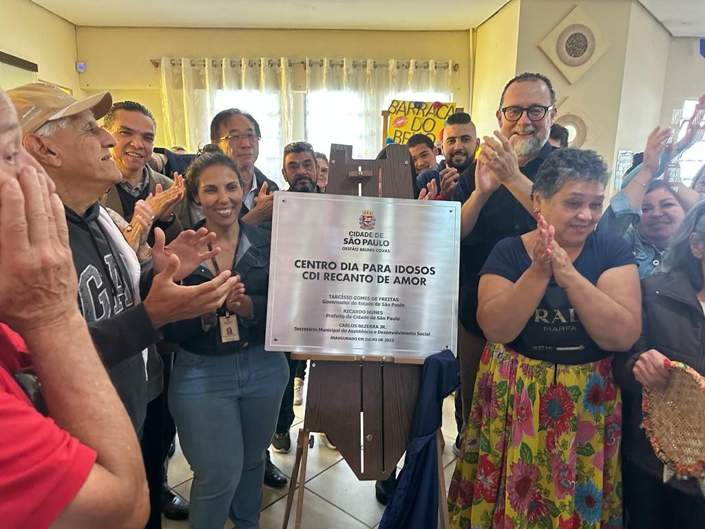 Foto que tem como destaque, no centro dela, a placa de inauguração do Centro Dia para Idosos Recanto de Amor. Ao redor dela, pessoas aplaudem.