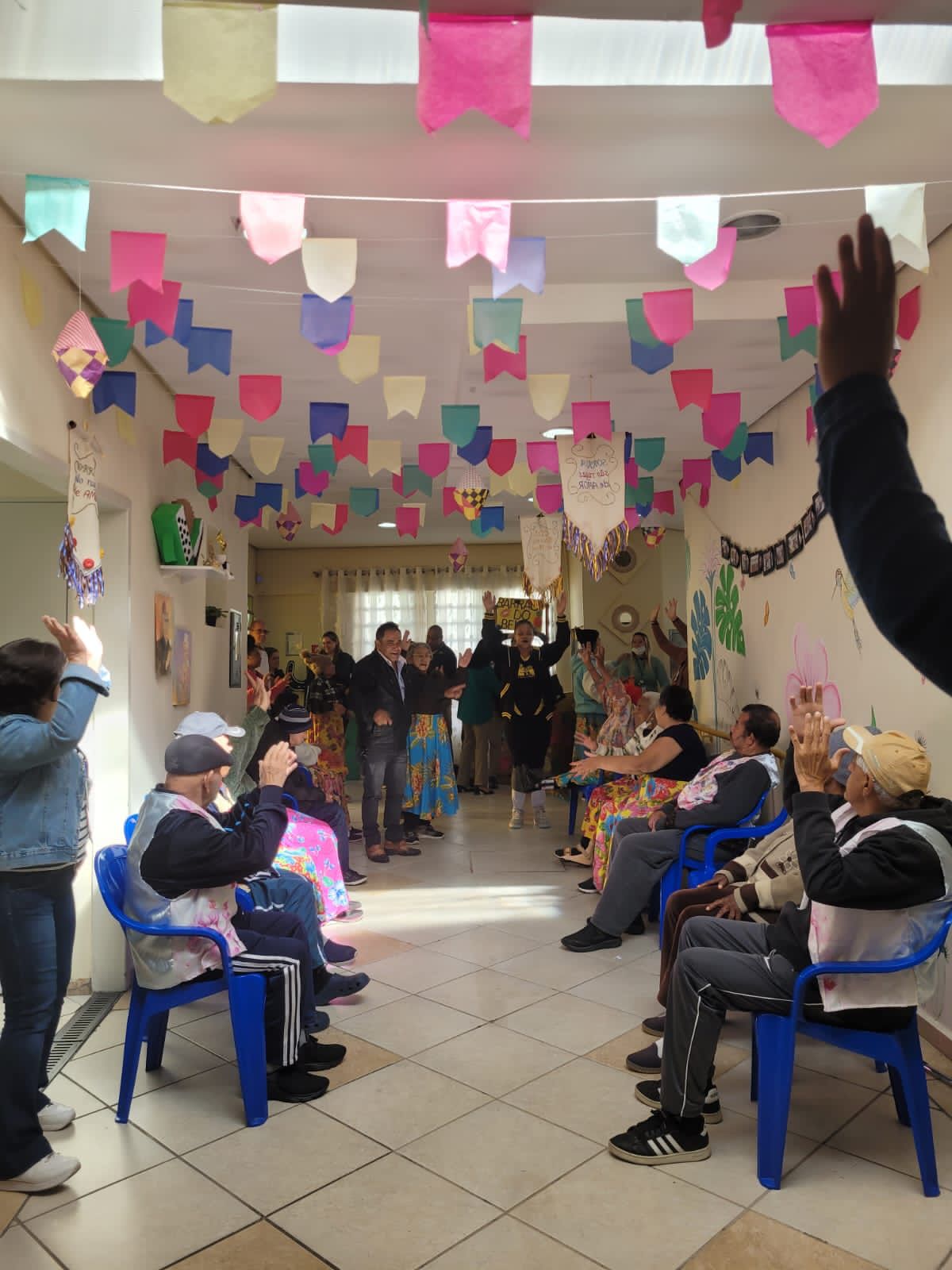 Foto de um corredor dentro do Centro Dia para Idosos Recanto de Amor durante uma festa. O ambiente está decorado com bandeiras coloridas típicas de festa junina e diversos detalhes nas paredes. Há varias pessoas na imagem participando da celebração.