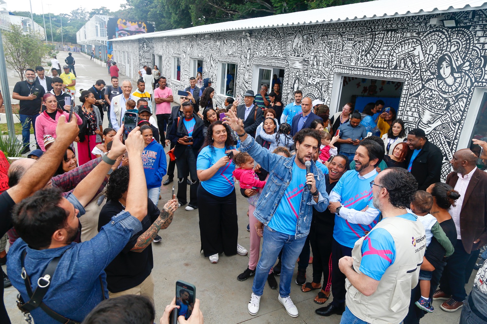 Na foto, diversos moradores e convidados estão reunidos assistindo uma orquestra. O prefeito Ricardo Nunes está no meio, ao lado do tenor Thiago Arancam. Eles cantam juntos.