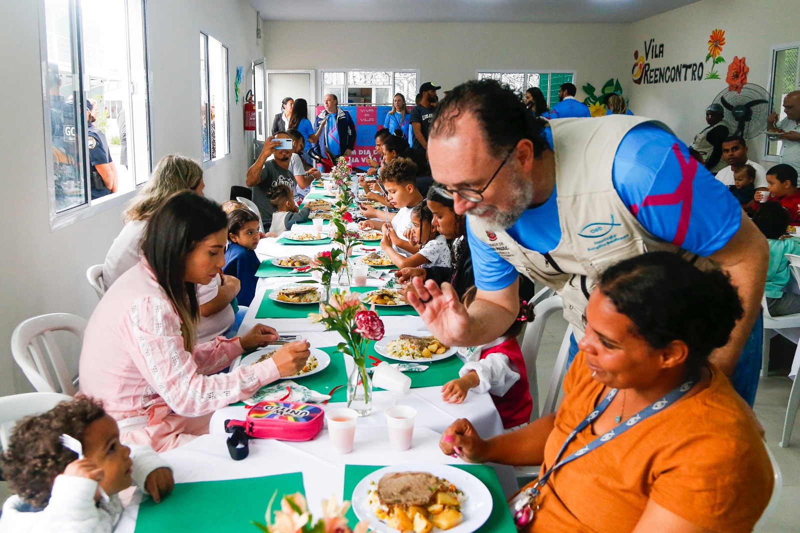 Na imagem, os moradores da Vila Reencontro Pari estão sentados em mesas brancas e almoçando. No fundo, há parede brancas e janelas em vidro. Atrás de uma das acolhidas, o secretário Carlos Bezerra Jr., inicia uma conversa.