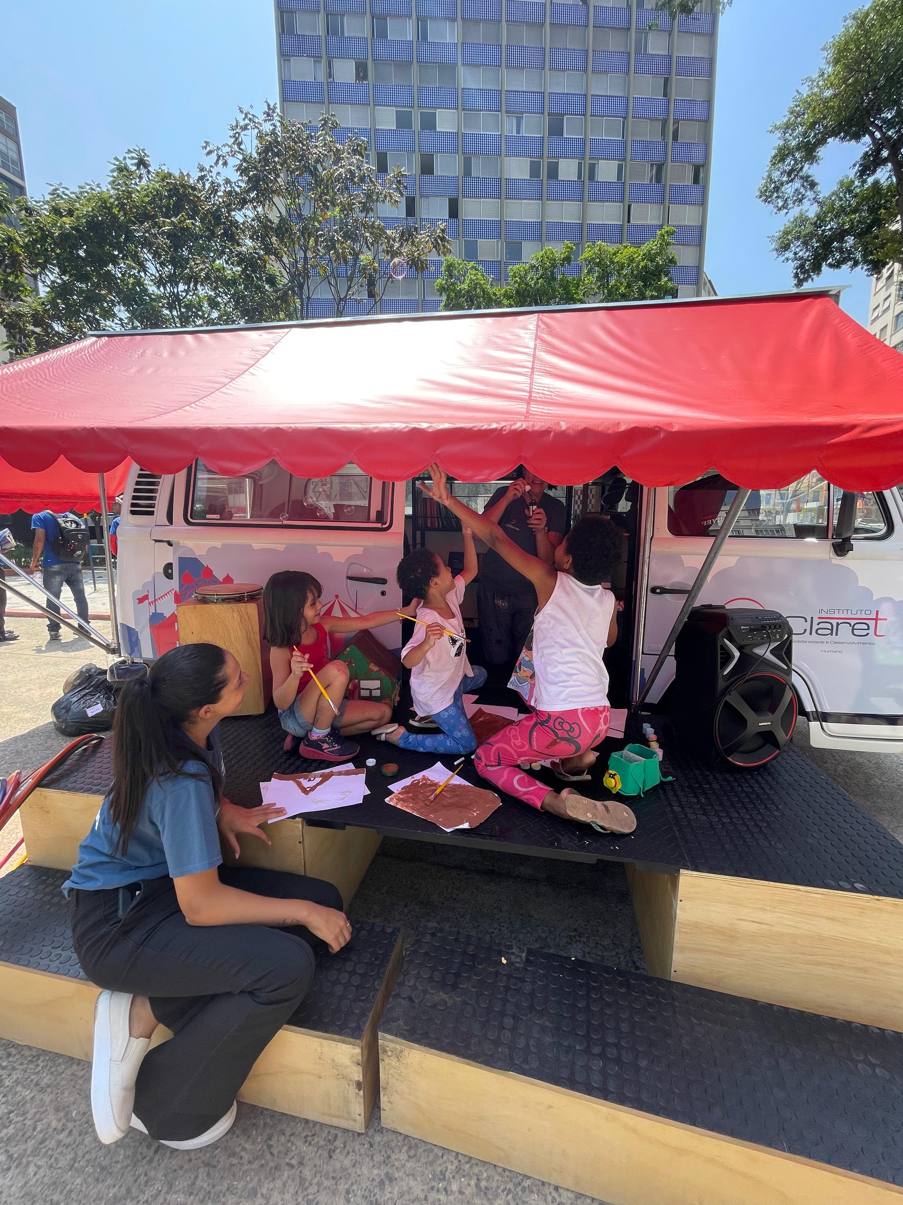 Crianças interagem com atividade promovida na Kombi do programa. Elas estão em cima de um pequeno palco, sob um toldo vermelho, observadas por uma agente social.