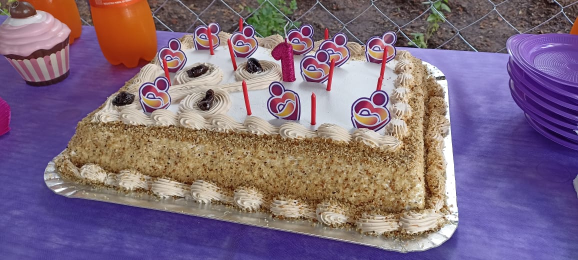 Imagem do bolo de aniversário da Vila Reencontro Anhangabaú. Há velas vermelhas e enfeites com o logo do Programa Reencontro sobre o bolo, além de uma vela rosa com o número um.