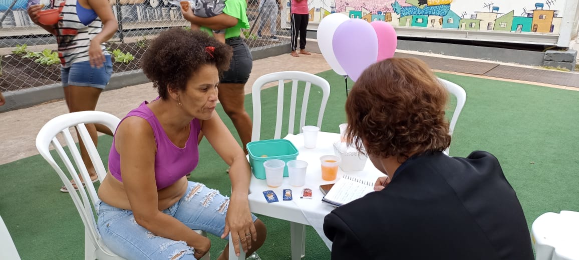 Imagem da acolhida Katia Semeão sentada à mesa enquanto cede entrevista. Ela veste uma blusa rosa e uma calça jeans azul. Há três bexigas coloridas de decoração compondo a foto.