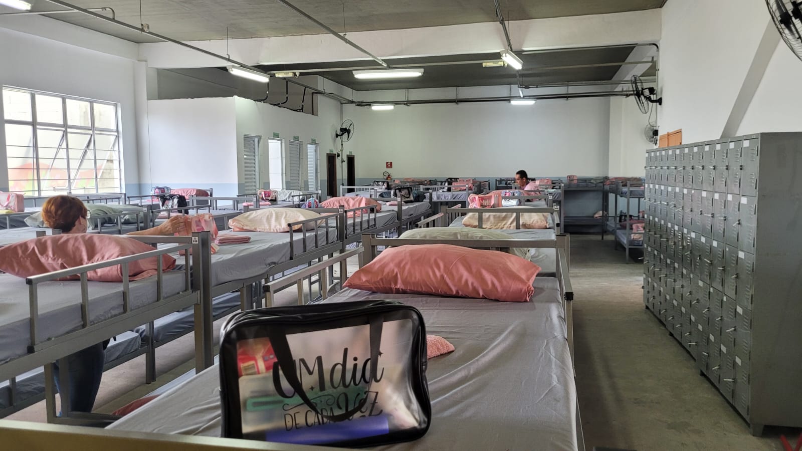 imagem de um dos quartos do Centro de Acolhida para Mulheres Lila Covas. Várias beliches estão dispostos. As camas estão forradas com lençol e têm um travesseiro e uma bolsa com objetos de higiene pessoal em cada uma delas.