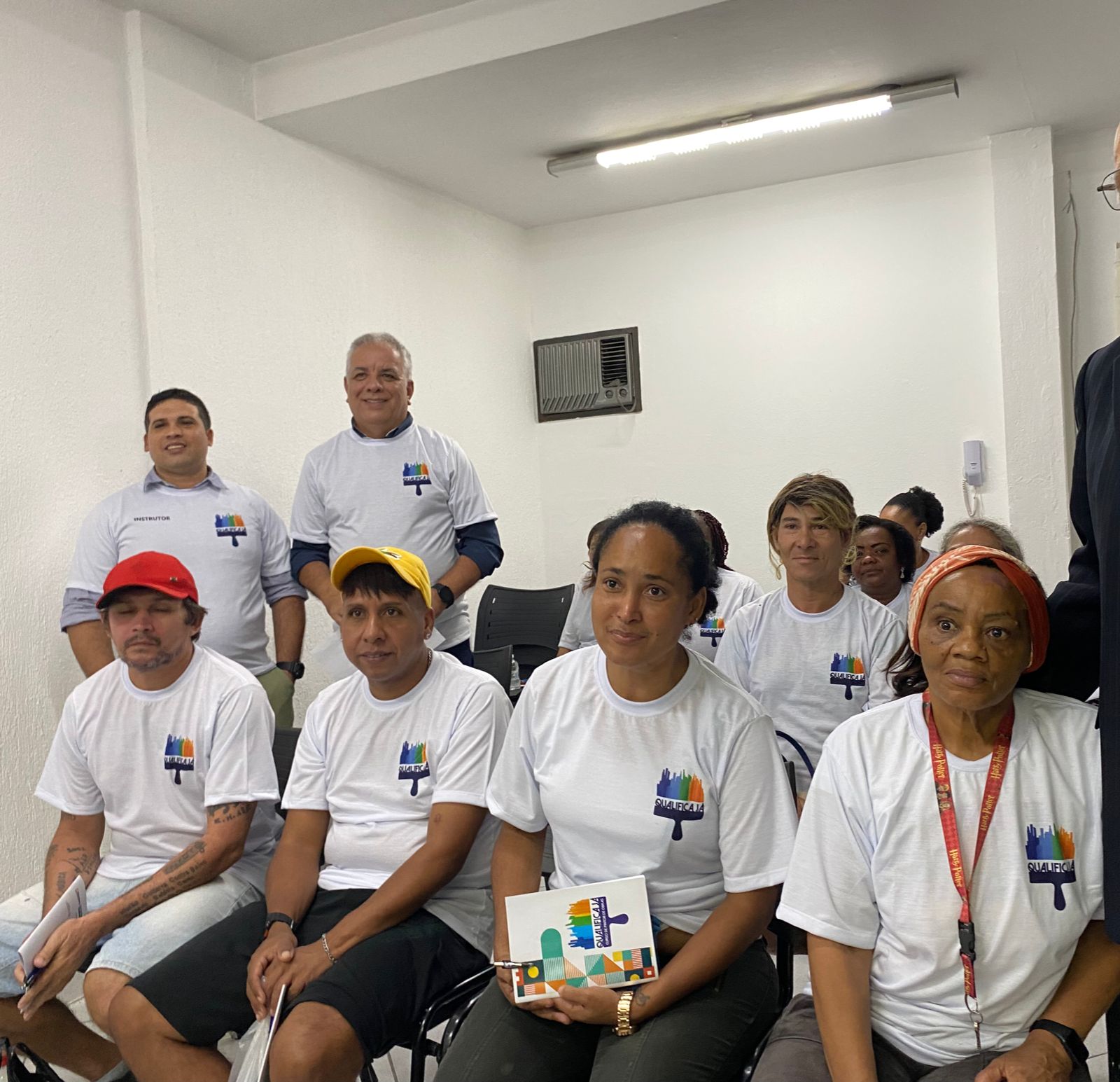 Alunos da primeira aula do Qualifica Já, alguns em pé e outros sentados, prestando atenção no conteúdo compartilhado. Todos trajam um uniforme branco.