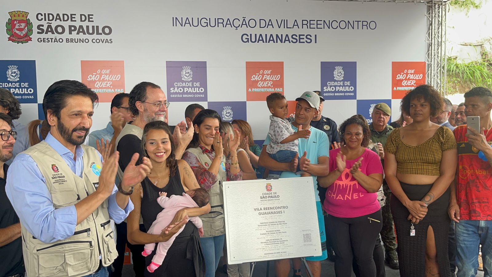 Diversas pessoas sorriem e posam para a foto atrás da placa de inauguração da Vila Reencontro Guaianases