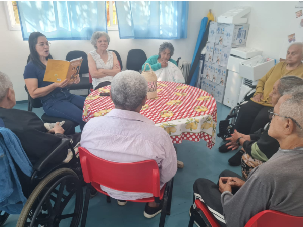 Na foto, diversos idosos estão reunidos para uma roda de conversas com café. a orientadora do serviço explica a atividade e segura um livro.