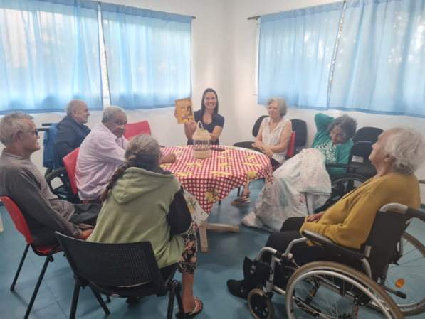 Na foto, diversos idosos estão reunidos para uma roda de conversas com café. a orientadora do serviço explica a atividade e segura um livro.