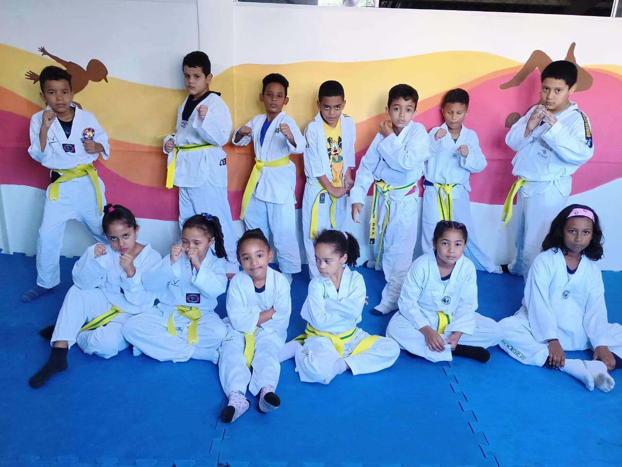 Na imagem, grupo de crianças com kimono branco e faixa amarela na cintura, elas estão em cima de um tatame.