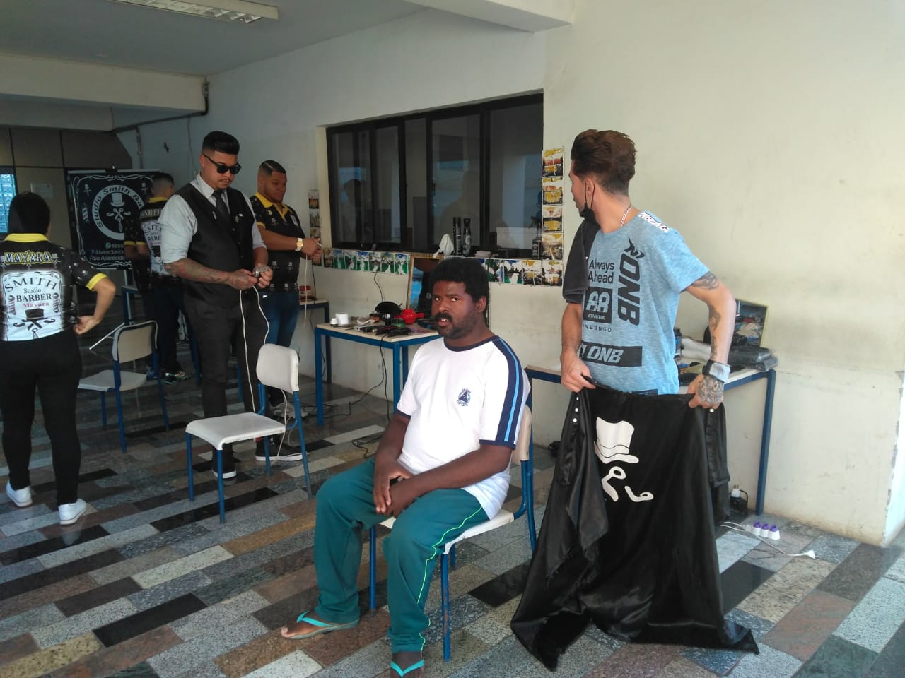 Homem de camiseta branca sentado em cadeira sendo atendido por barbeiro; ao fundo outros quatro profissionais fazem manutenção em seus equipamentos.