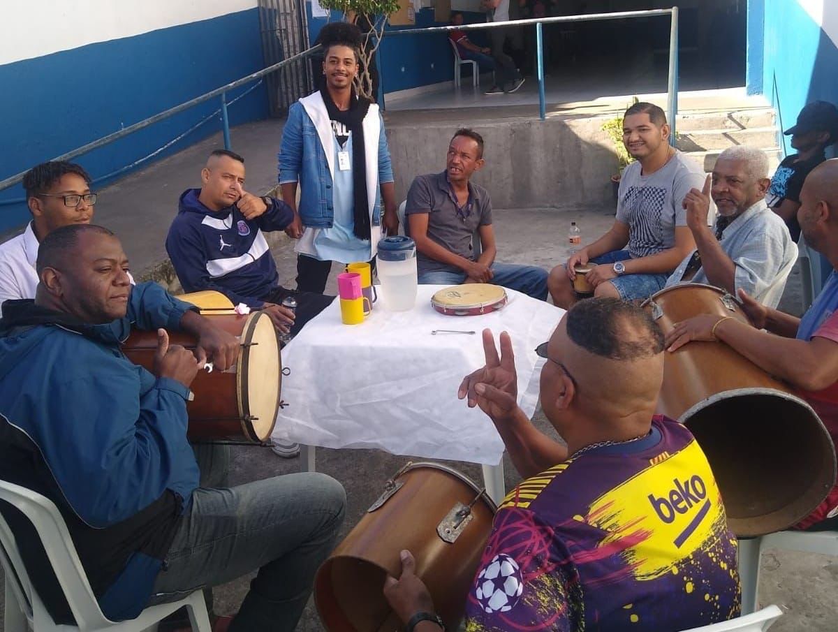 Mãos de orientadora tocando um instrumento de corda. No fundo, três atendidos tocam tambor e pandeiro, e estão em frente a um portão.