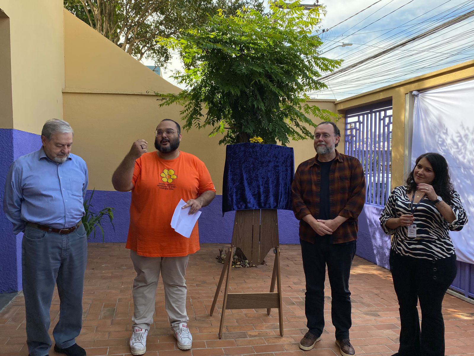 Equipe do serviço dircursa ao lado do secretário durante inauguração. No meio, um cavalete marrom sustenta a placa do SPVV, que estã coberta por um pano azul.