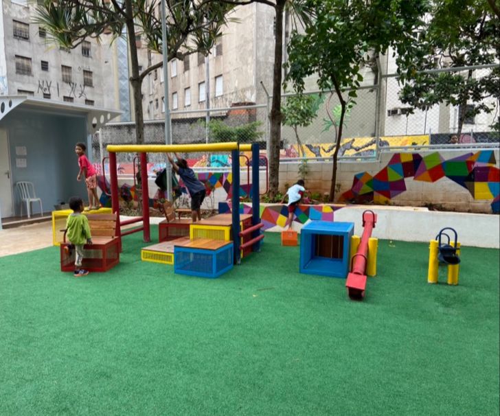 Na foto um playground na Vila Reencontro. Os brinquedos são coloridos e estão postos em um gramado sintético. No fundo uma parede abstrata colorida e prédios em cinza