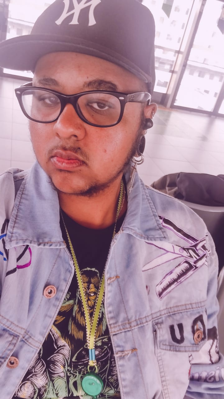 Homem usando jaqueta jeans e boné olha para camera. O homem está usando óculos e possui barba no queijo.
