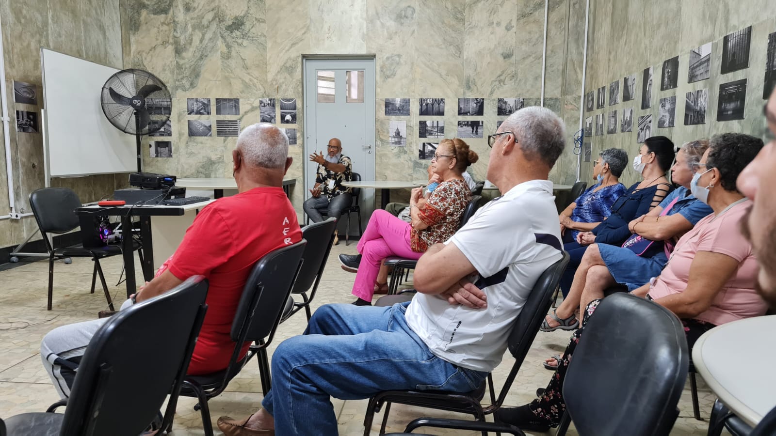 Na foto os conviventes estão sentados em cadeiras pretas enquanto acompanham a palestra. No fundo, fotos estão coladas nas paredes 