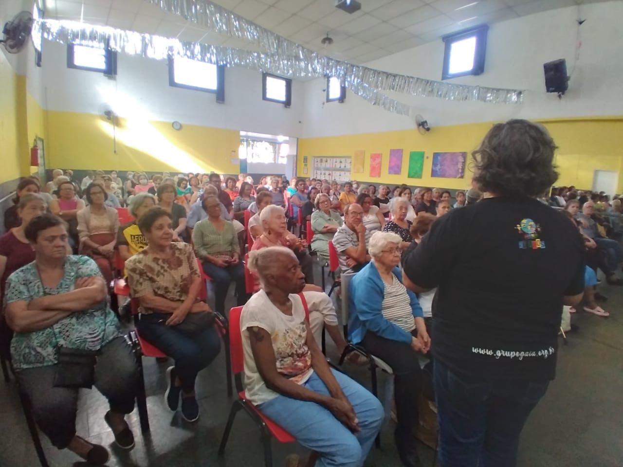 Atendidos do GAIA prestigiando palestra sobre imigração. No teto branco, varais com fitas prateadas decoram o espaço. As paredes da sala são amarelas. Há algumas artes coladas e cinco janelas.