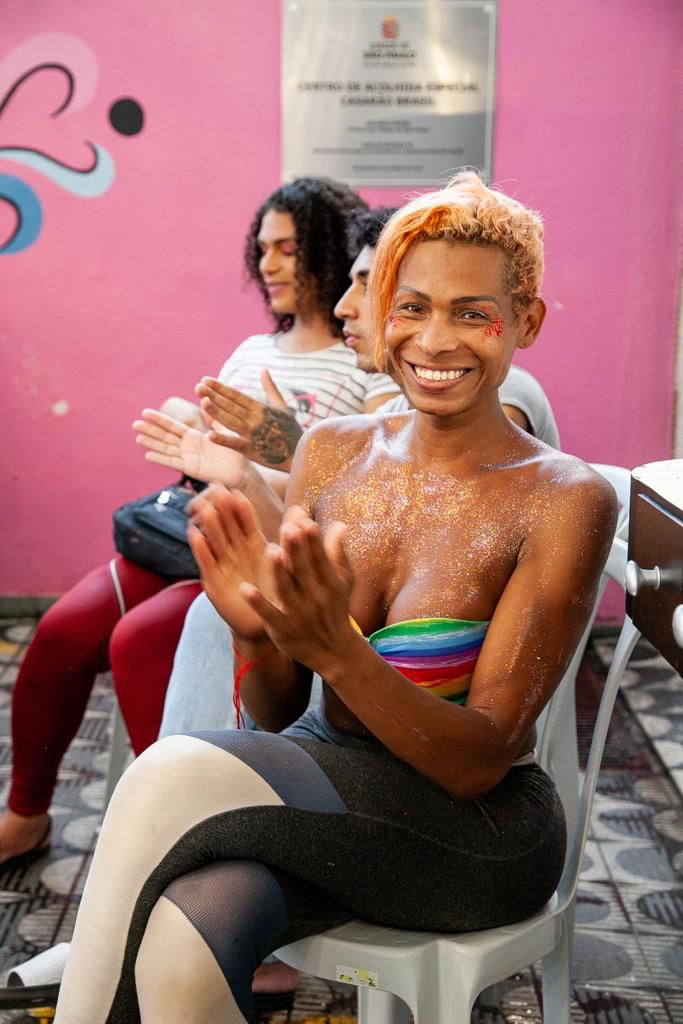 Sentada em uma cadeira de plástico, uma mulher com cabelos alaranjados, vestindo um sutiã colorido e uma calça leg preta com detalhes brancos, posa para foto enquanto bate palmas.