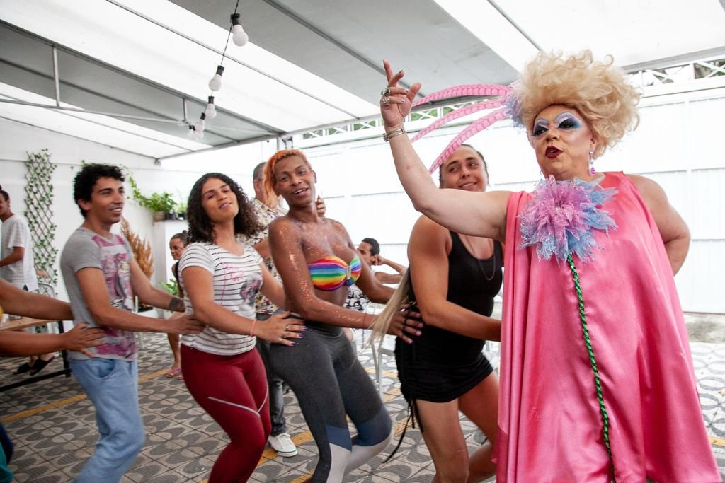 Drag queen vestindo um vestido cor de rosa, com os cabelos loiros e armados, está dançando com outras mulheres que formam uma fila atrás dela.