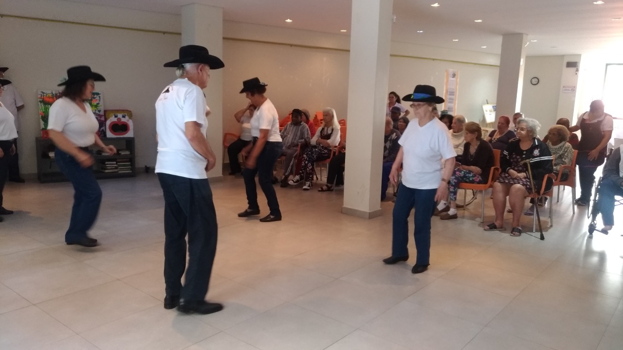 Cinco idosos estão de pé, usando camisetas brancas, calças jeans e chapéus. Enquanto eles se apresentam, outros idosos, sentados em cadeiras laranjas, observam a apresentação.