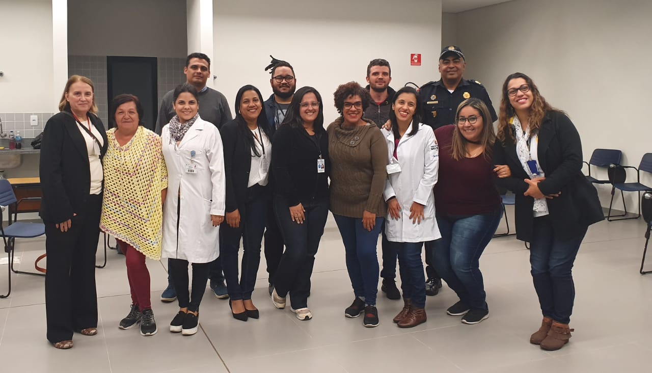 Nove mulheres e quatro homens estão pé, dentro de uma sala e olhando para a foto. As mulheres ficam a frente e os homens atrás. Ao redor deles, há cadeiras espalhadas.