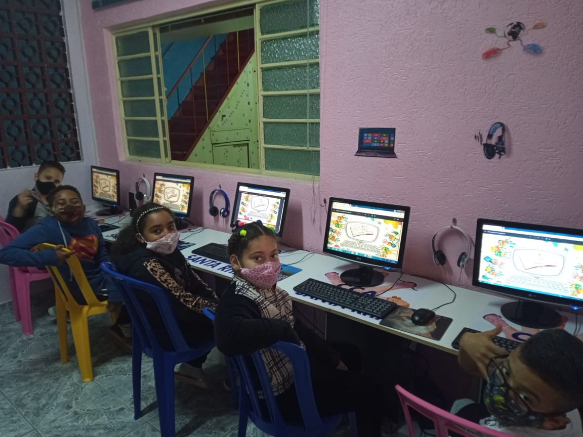 Foto da sala de informática do CCA com cinco computadores e cinco crianças que estão acessando a biblioteca virtual
