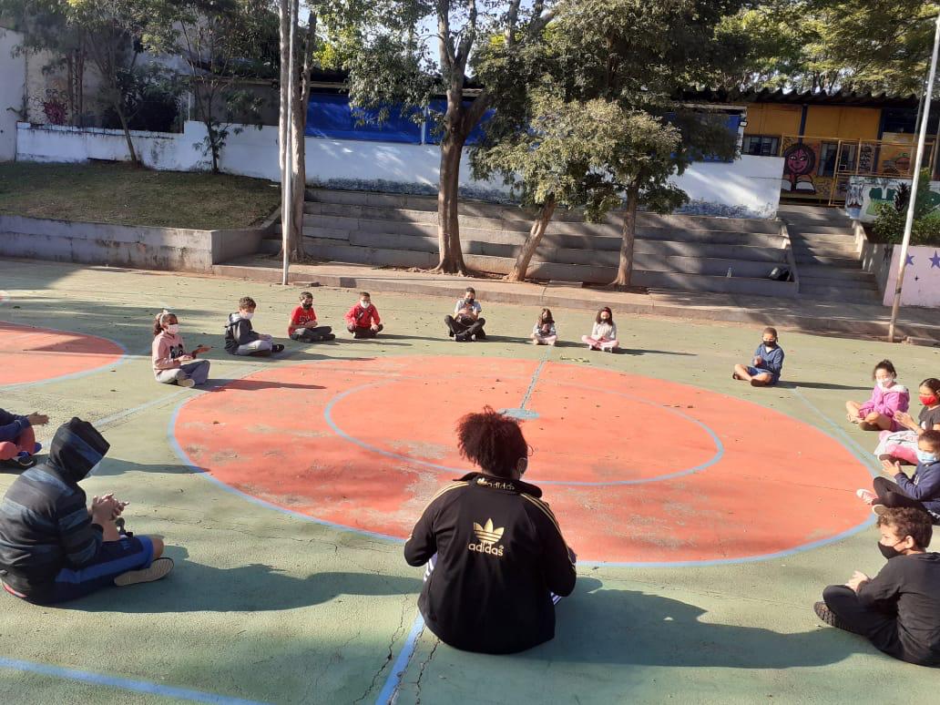 Várias crianças estão sentadas em um grande círculo na quadra esportiva do serviço. Todas estão usando máscara de proteção facial e há uma profissional orientando a atividade. 