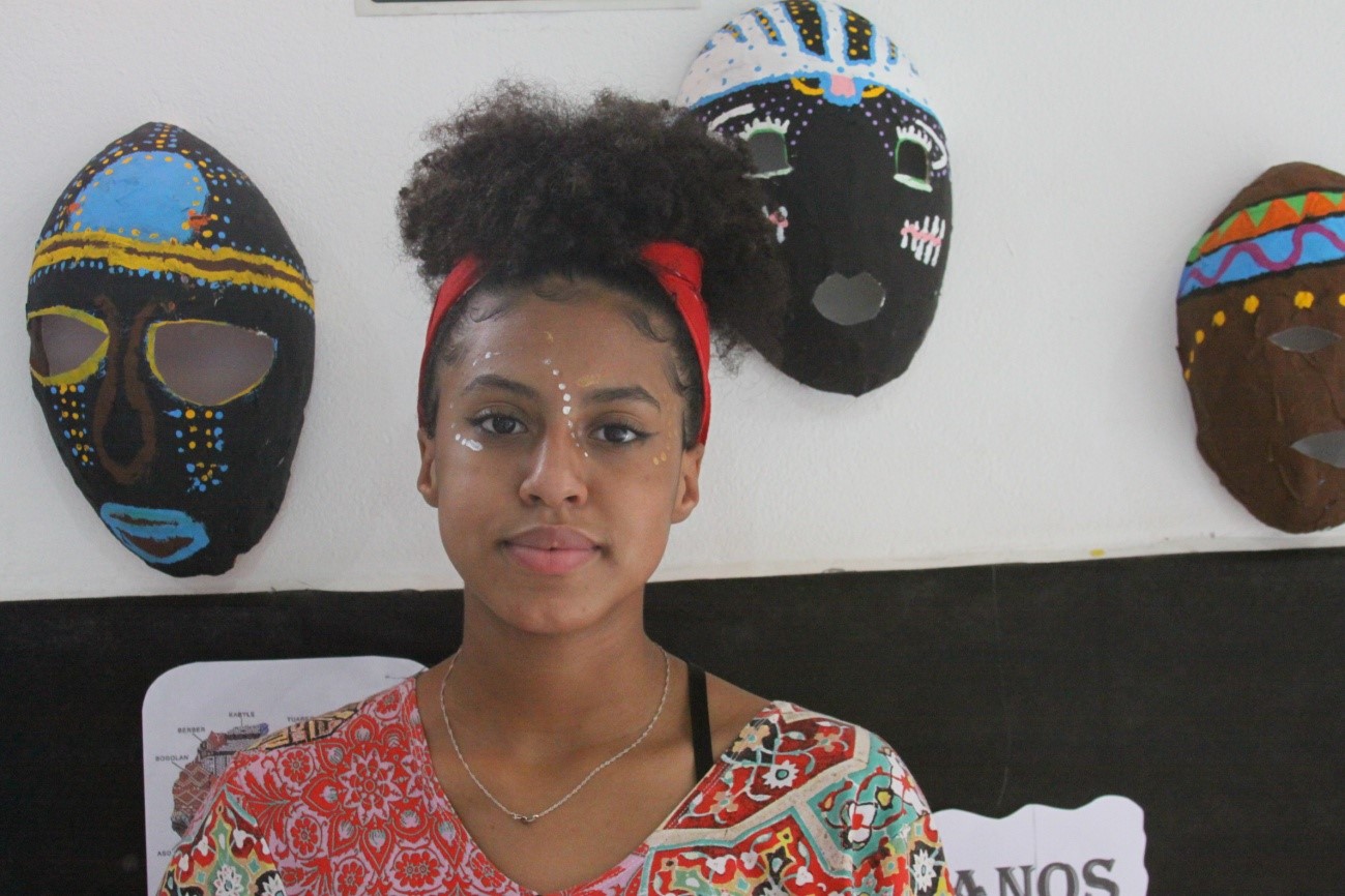 Convivente do serviço com maquiagem afro, logo atrás mascaras afros penduradas na parede.