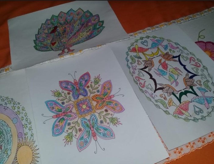 Sobre uma mesa com toalha laranja estão três folhas com desenhos impressos pintados pelos idosos com lápis de cor. O desenho da folha central é de borboletas e o das outras duas folhas são de pássaros. 