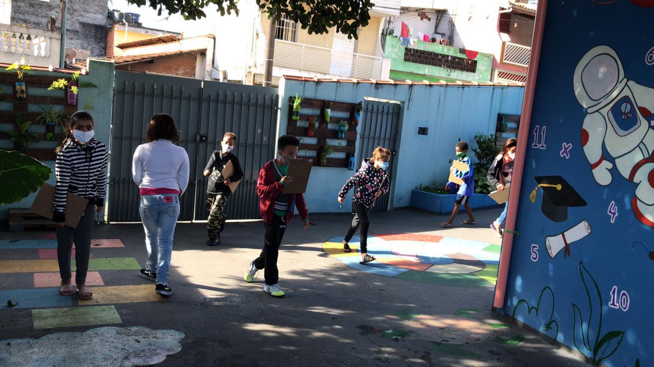 Devidamente adequadas aos protocolos sanitários, crianças na área externa do CCA estão com pranchetas na mão, realizando atividade da oficina.