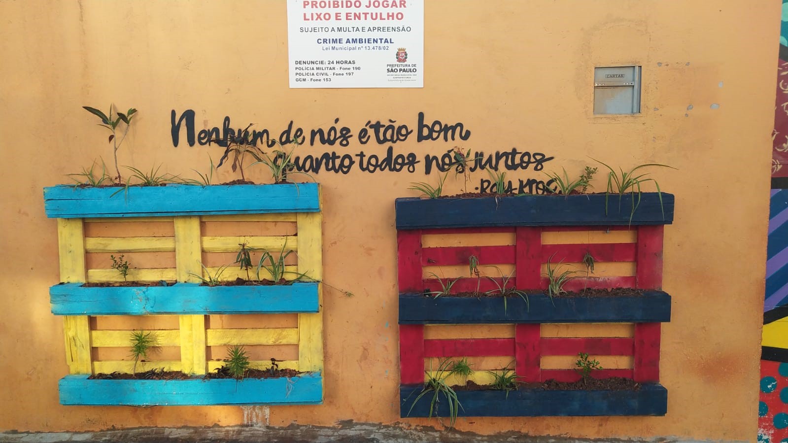 Parede amarelado com uma placa pendurada escrito “proibido jogar lixo e entulho” Embaixo da placa tem uma frase “Nenhum de nós é tão bom quanto todos nós juntos” e abaixo dois paletes que são do jardim suspenso, o da esquerda é amarelo e azul e o da direita é vermelho e preto.
