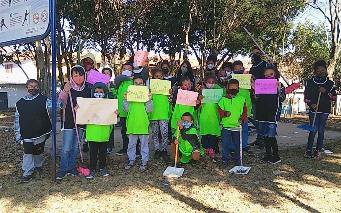 Vinte duas crianças e adolescentes de máscara reunidas em uma praça. Algumas delas seguram cartazes e outros utensílios de limpeza.
