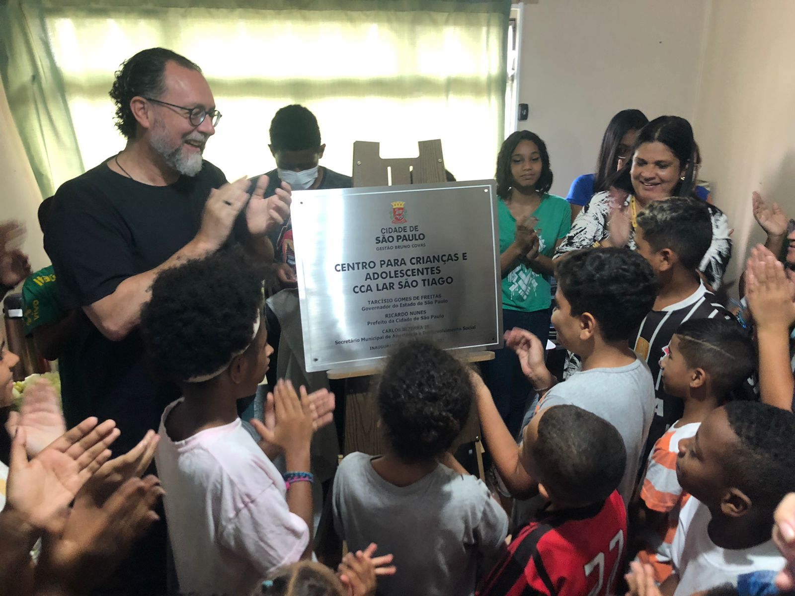 Na foto, adultos e crianças estão ao lado e em frente do secretário Carlos Bezerra aplaudindo a inauguração do CCA. No centro tem um cavalete com a placa de identificação do serviço em cima.