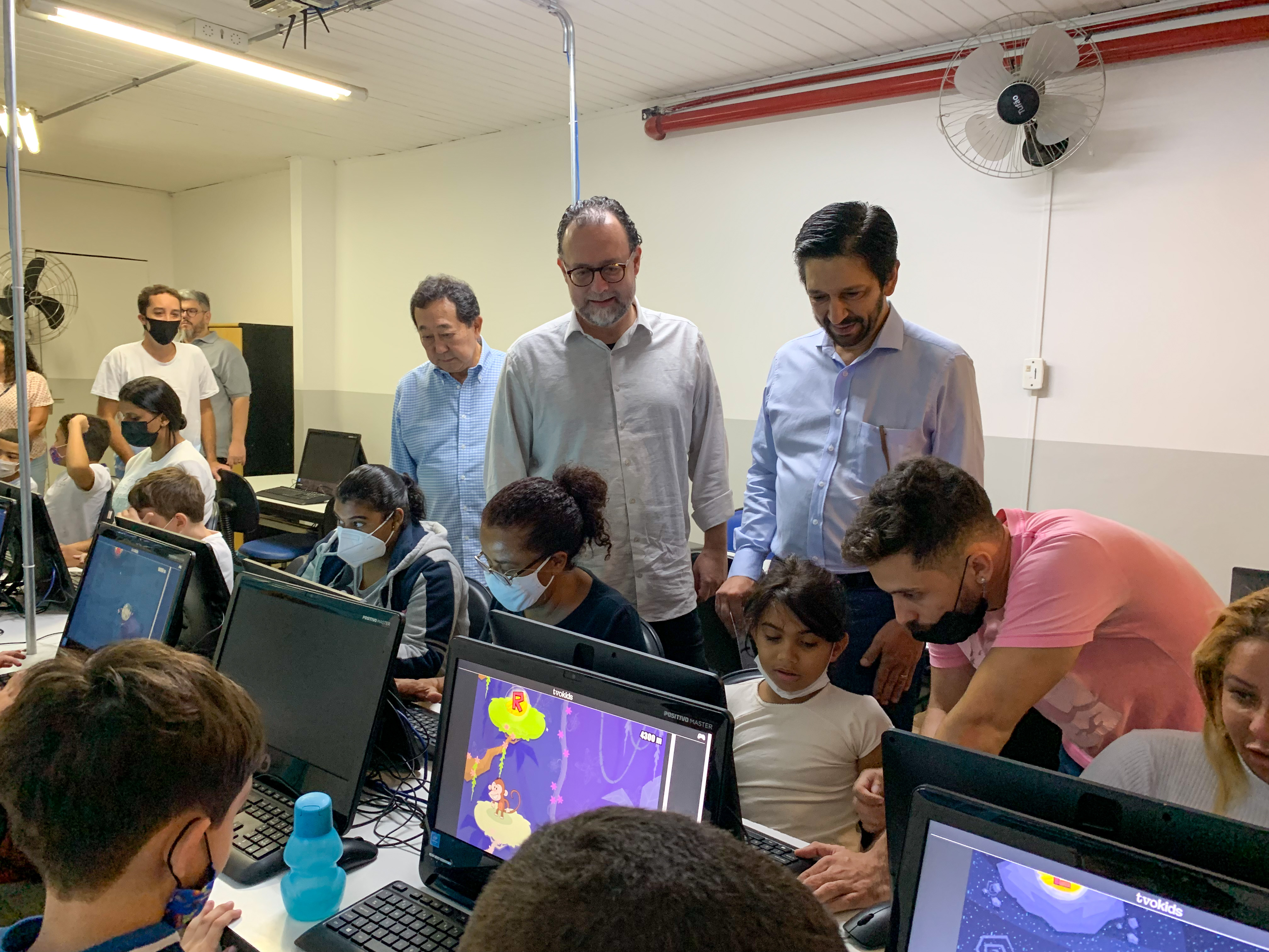 Secretario e Prefeito em pé vendo as crianças sentadas mexendo em computadores.