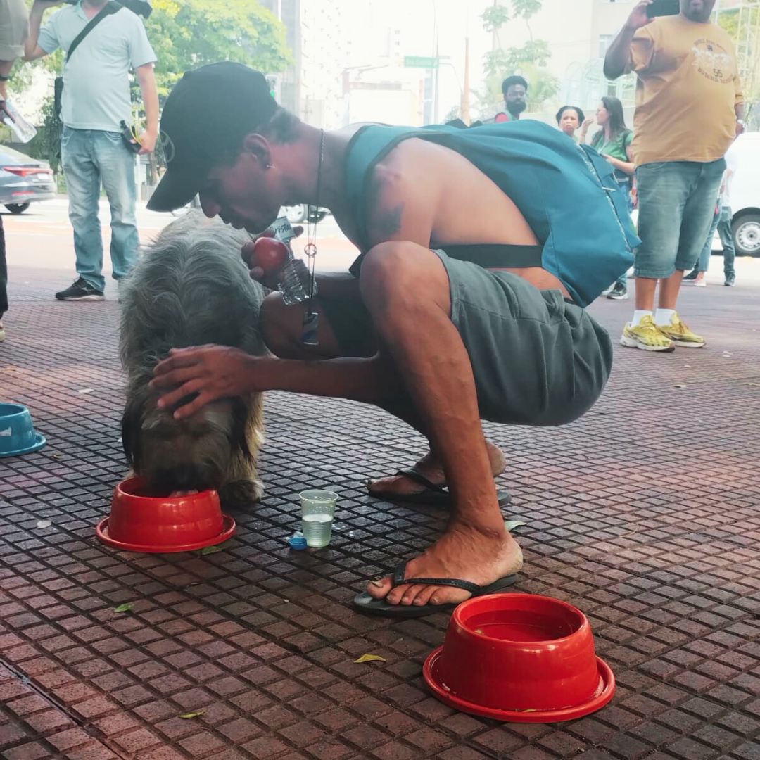 Na imagem há um cachorro de porte médio bebendo agua de um pote vermelho que está no chão. Ao lado dele um hoemem abaixado sem blusa, usando um shortes preto e bone preto