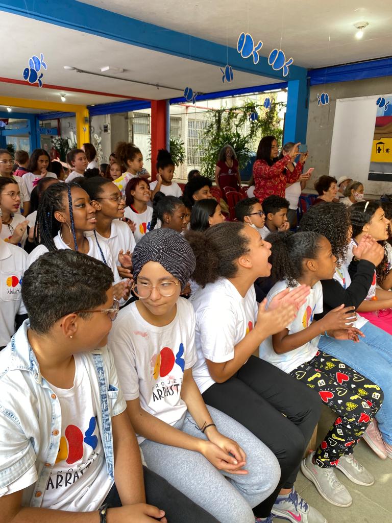 Diversas crianças sentadas em bancos, lado a lado, e assistindo apresentações.