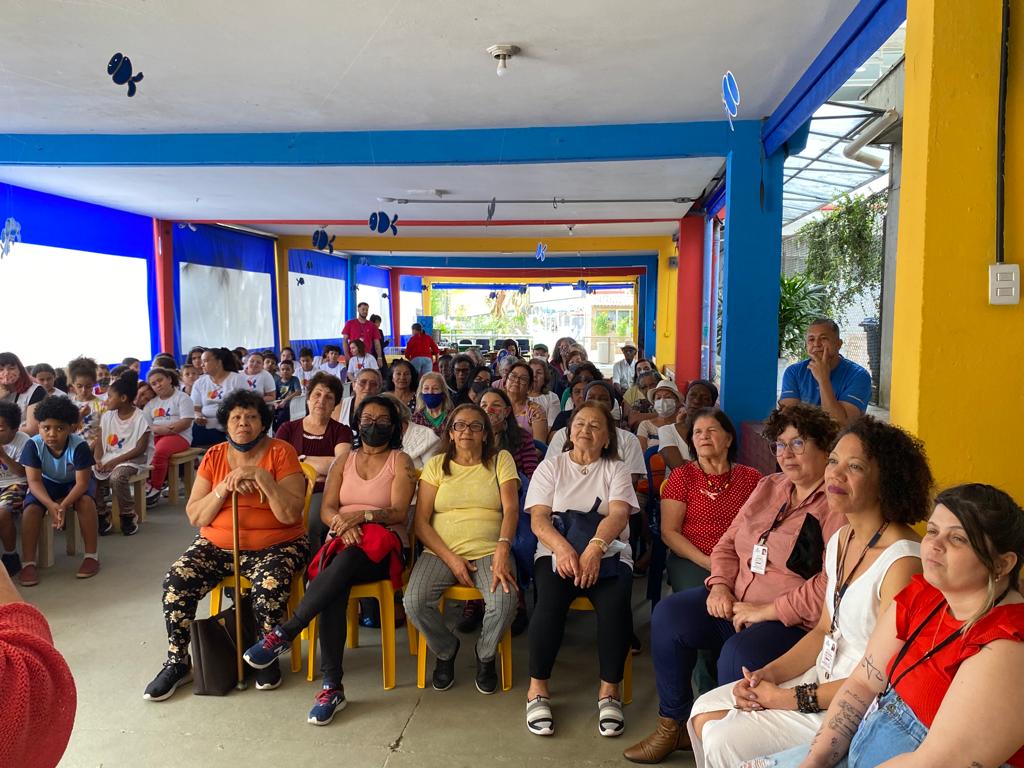 Mulheres sentadas a direita. Crianças sentadas a esquerda. Todos assistindo uma apresentação.