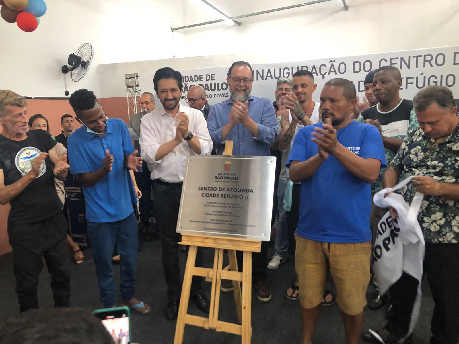 Na foto há oito homens ao redor de um cavalete batendo palmas e sorrindo. No meio, em cima do cavalete, está uma placa de metal escrita ‘Centro de Acolhida Cidade Refúgio II’.