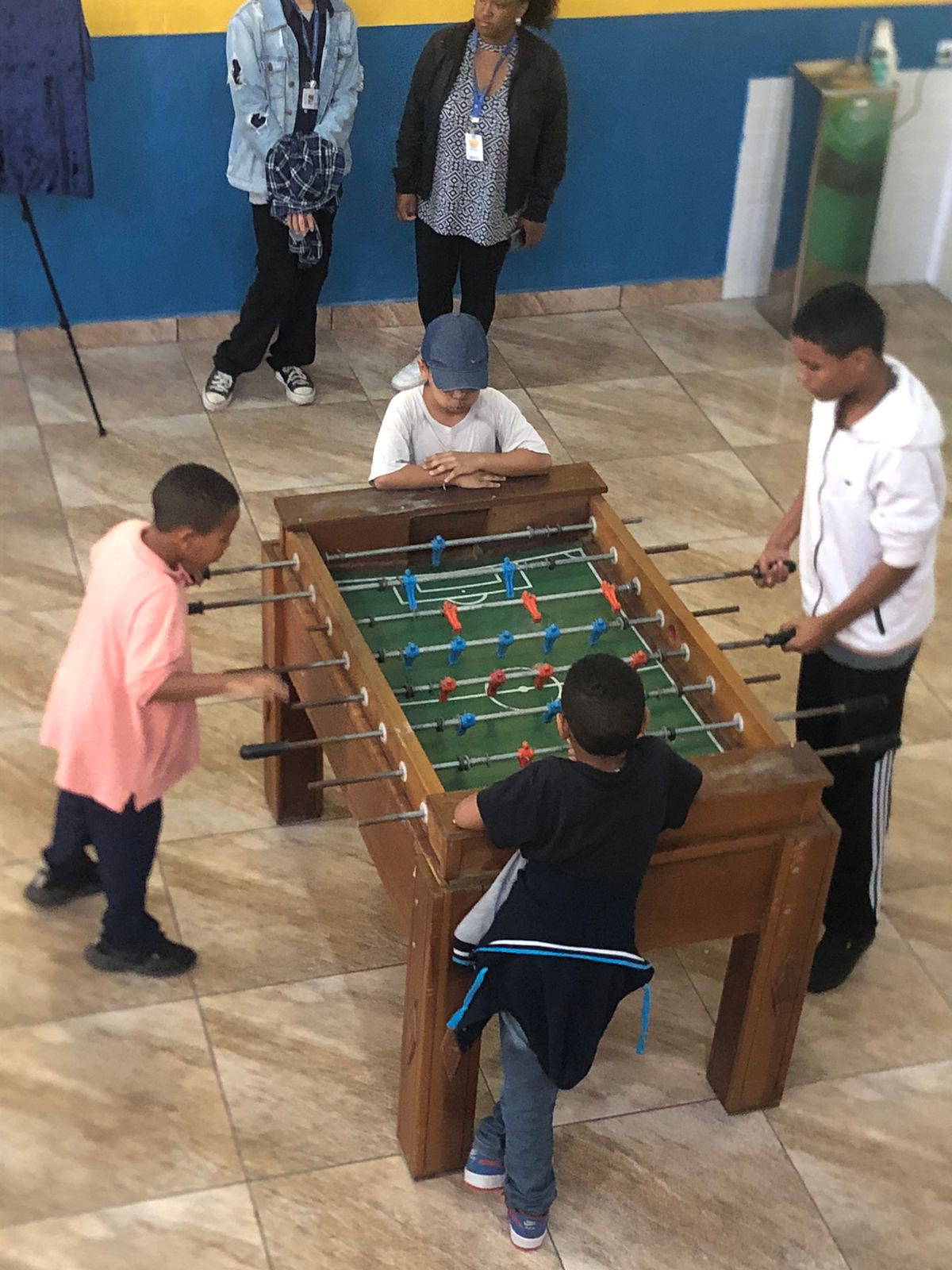 Dois meninos estão brincando em uma mesa de pebolim. Outros dois acompanham o jogo nas duas pontas da mesa. 