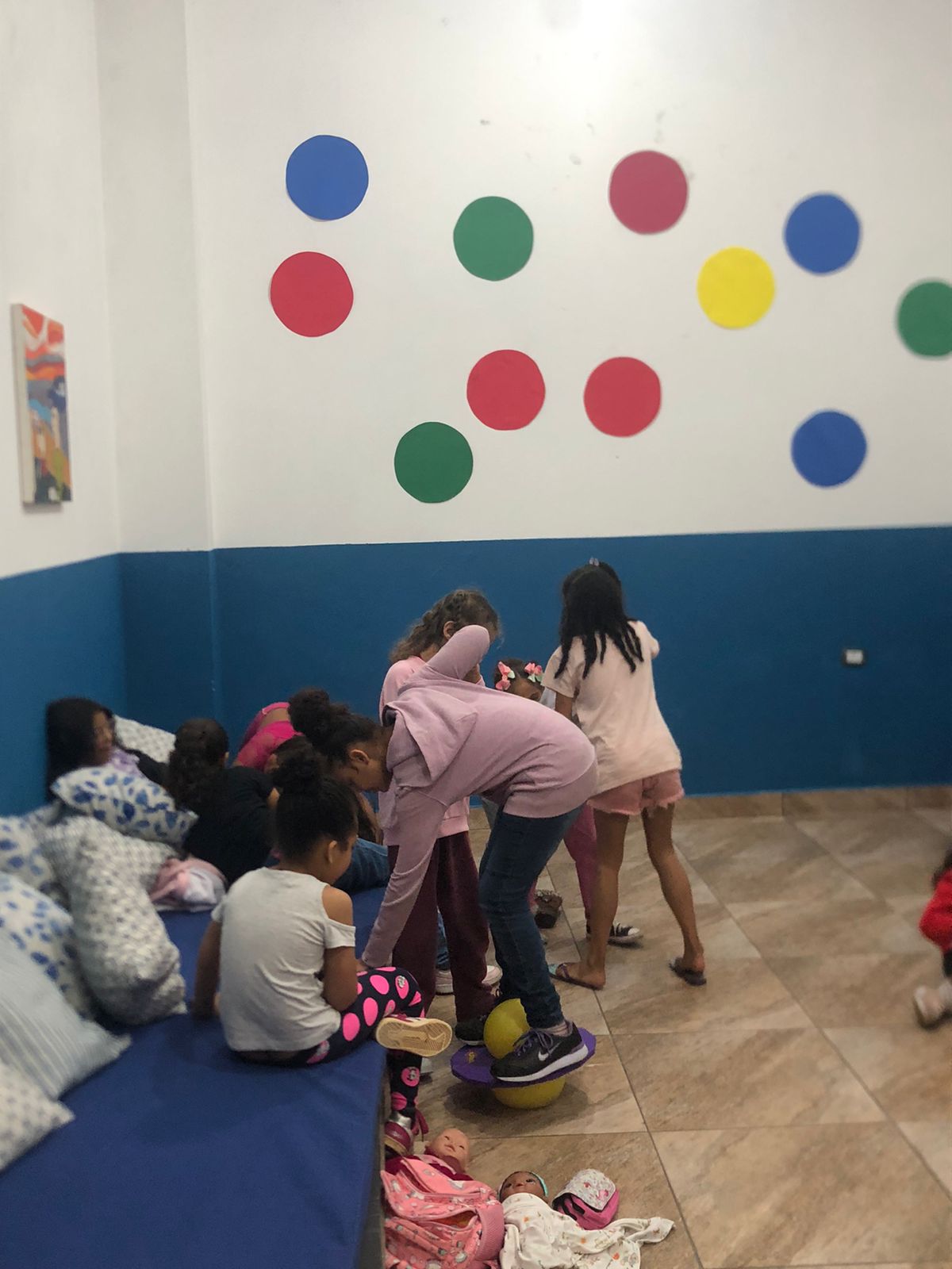 Algumas meninas estão brincando no espaço da brinquedoteca do serviço. Algumas estão em pé e outras sentadas em um sofá. No fundo, há uma parede pintada com as cores azul escuro e branco, e, dividida nas mesmas cores, respectivamente. Algumas esferas coloridas foram coladas na parede. 