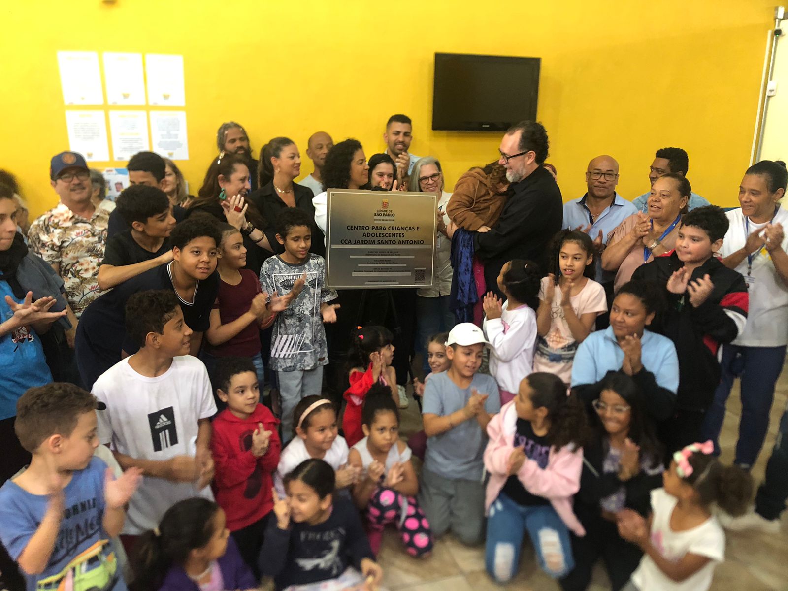 Crianças do Centro para Crianças e Adolescentes (CCA) ‘Jardim Santo Antônio’ estão em volta da placa de inauguração do serviço. O secretário Carlos Bezerra Jr. está sorrindo e segurando uma menina no colo. 