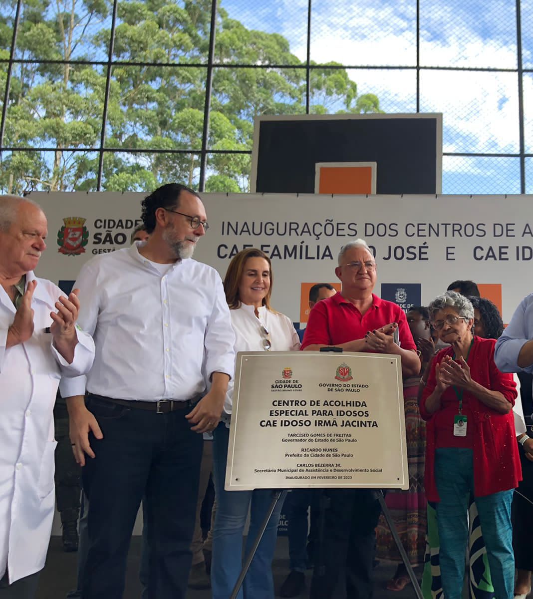 Quatro pessoas em pé apalaudindo. Em frente a elas, esta uma placa de metal escrito 'Centros de Acolhida Especial (CAE) para Idosos Irmã Jacinta’.