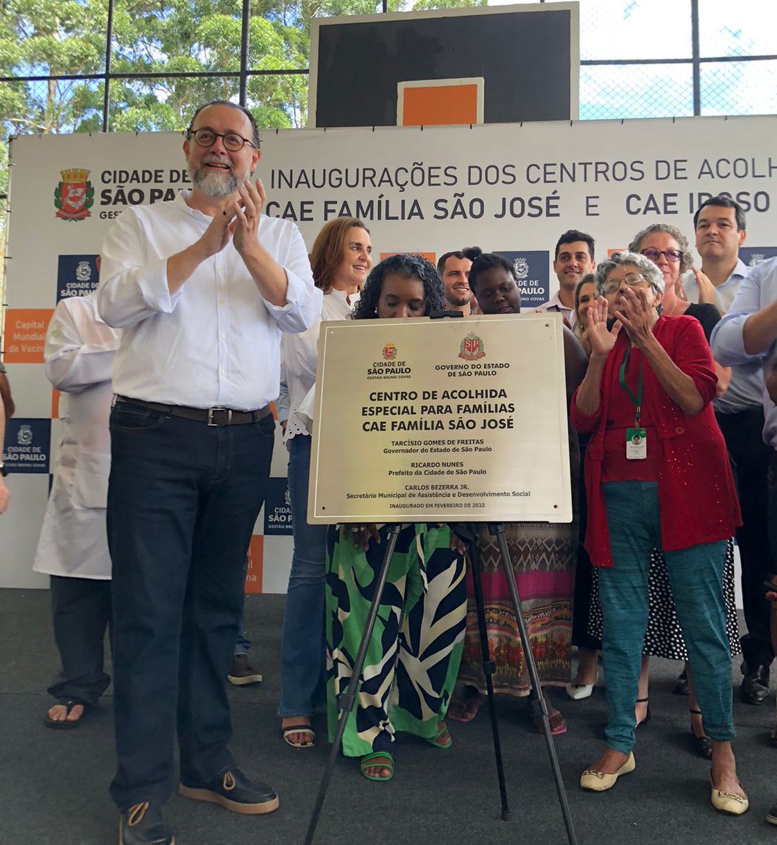 Dez pessoas em pé apalaudindo. Em frente a elas, esta uma placa de metal escrito 'Centros de Acolhida Especial (CAE) para Famílias São José’. 