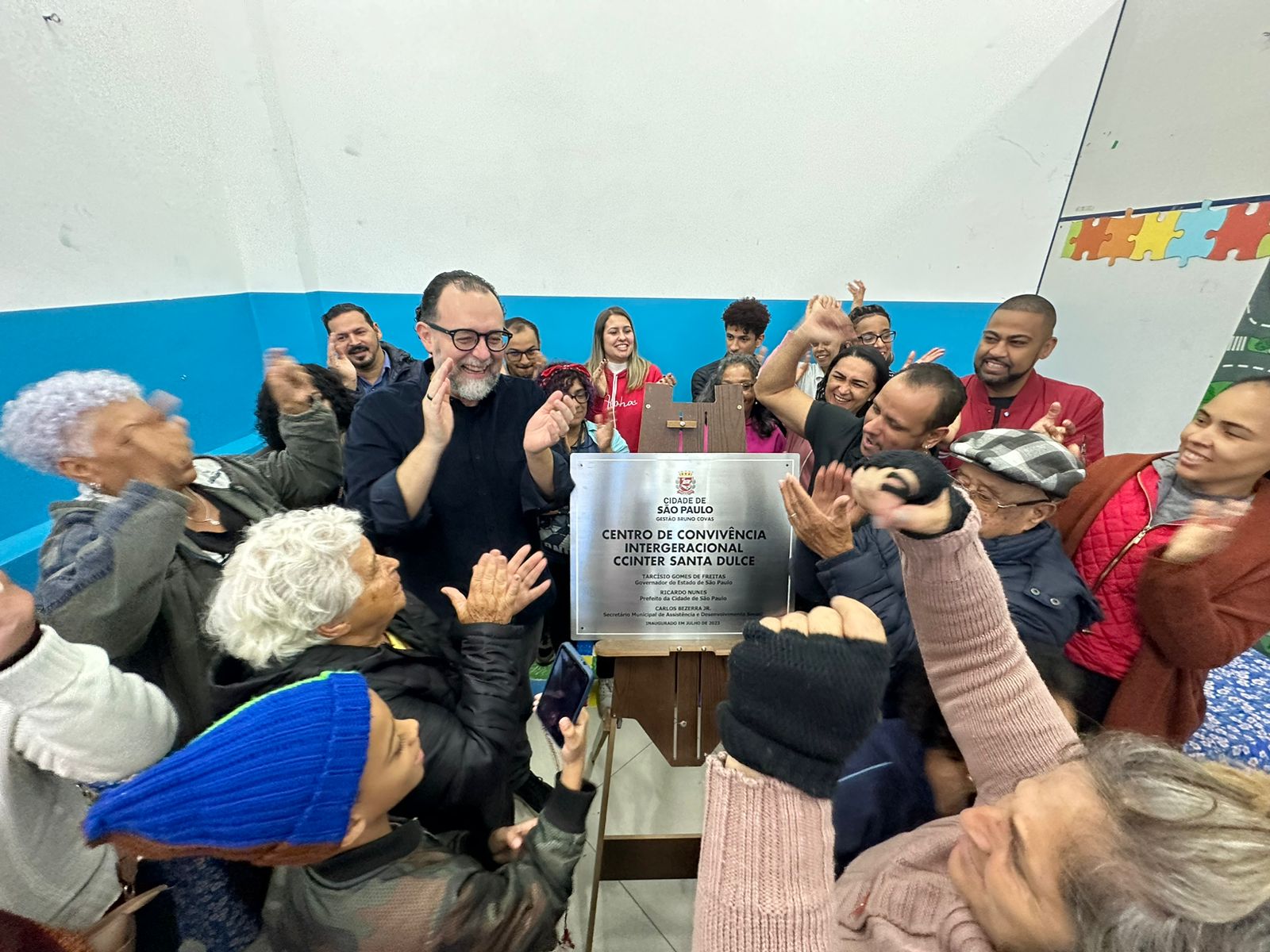 Pessoas em pé batendo palmas. No meio, cavalete de madeira, com uma placa de aço inox no meio escrito Centro de Convivência Intergeracional (CCInter) 'Santa Dulce'