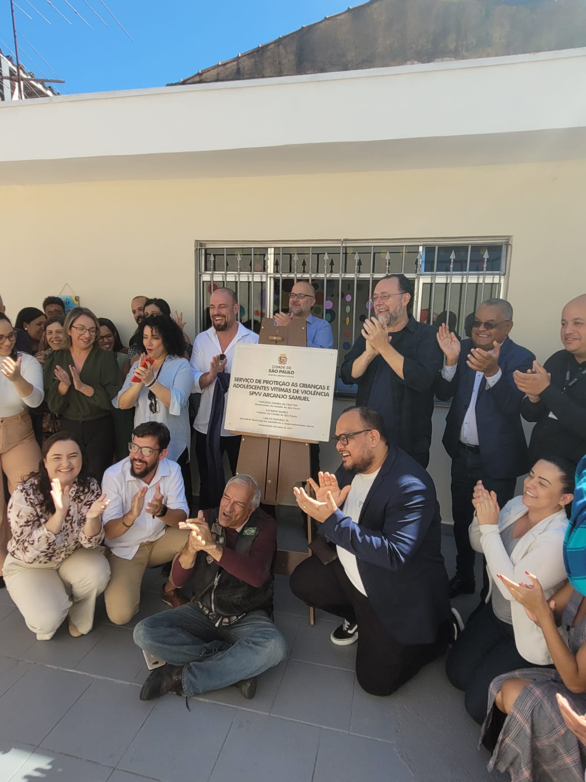 No centro da foto, a placa de inauguração do SPVV 'Arcanjo Samuel'. Ao redor da placa, o secretário de Assistência e Desenvolvimento Social, e as pessoas que participaram da cerimônia, sorrindo e aplaudindo.