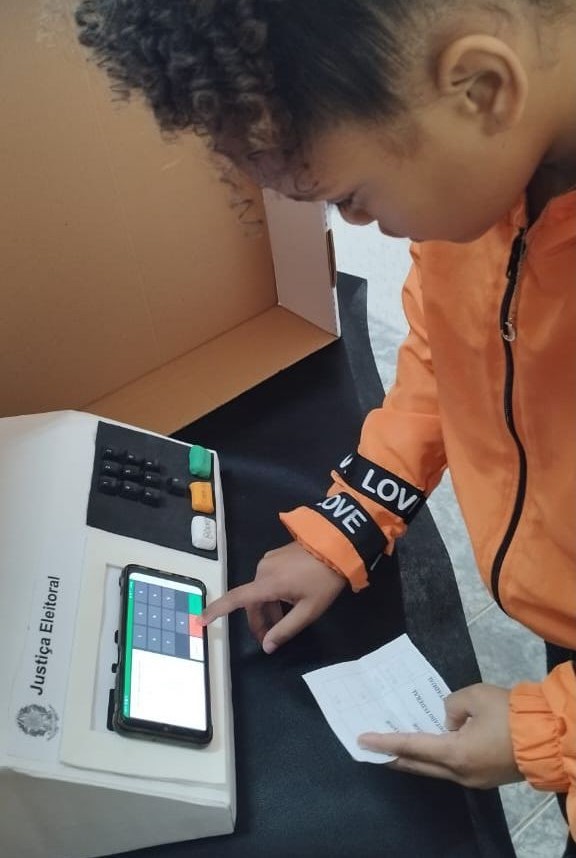 Uma menina com uma blusa de frio laranja com um papel na mão e digitando na urna que foi confeccionada pelos alunos. 