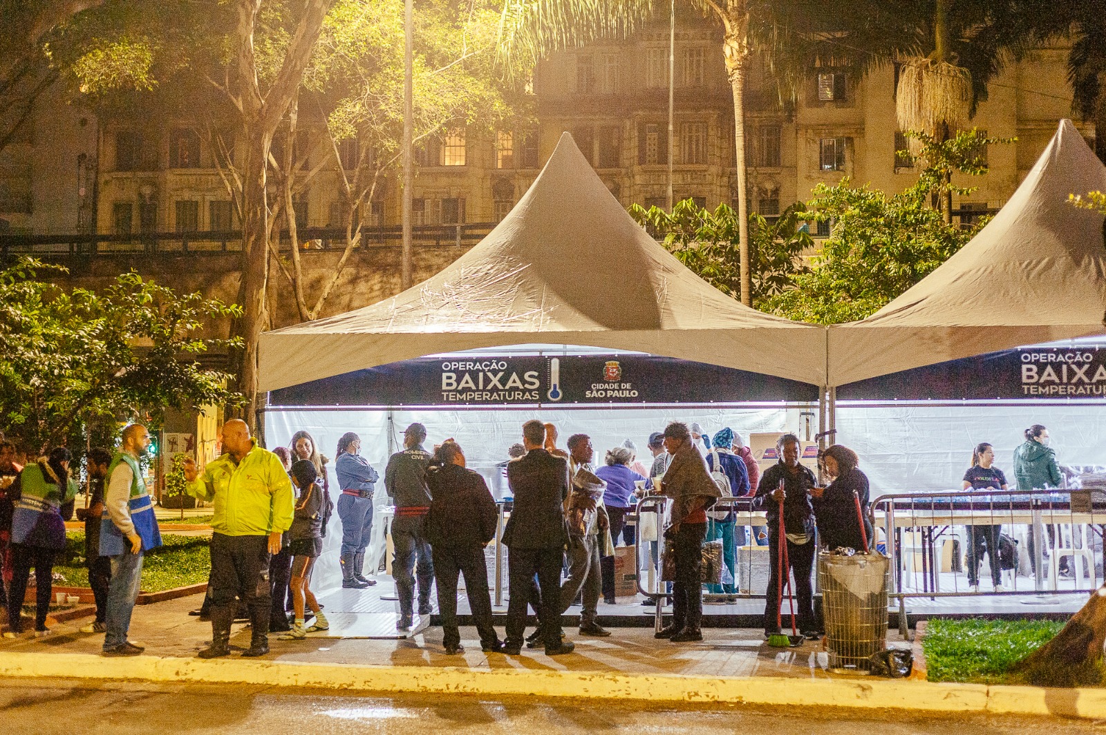  Foto em ambiente aberto. No meio, duas tendas montadas e um grupo grande de pessoas em pé na frente. Ao redor da tenda há muitas arvores e ao fundo da imagem há um prédio largo e alto.