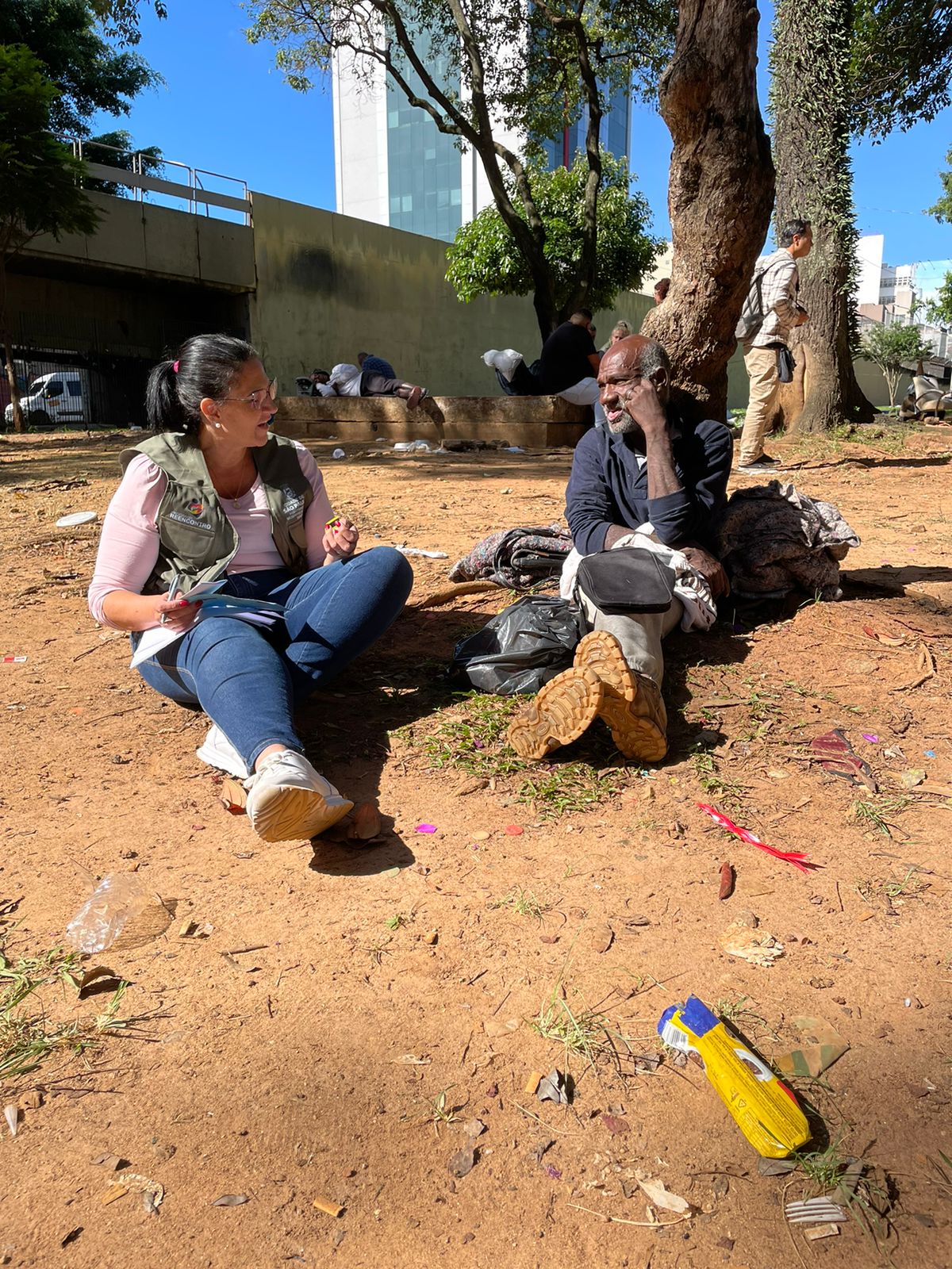 Atendimento social na Praça da Armênia. Duas pessoas sentadas na terra, sendo a profissional do Ampara SP e a pessoa em situação de rua.