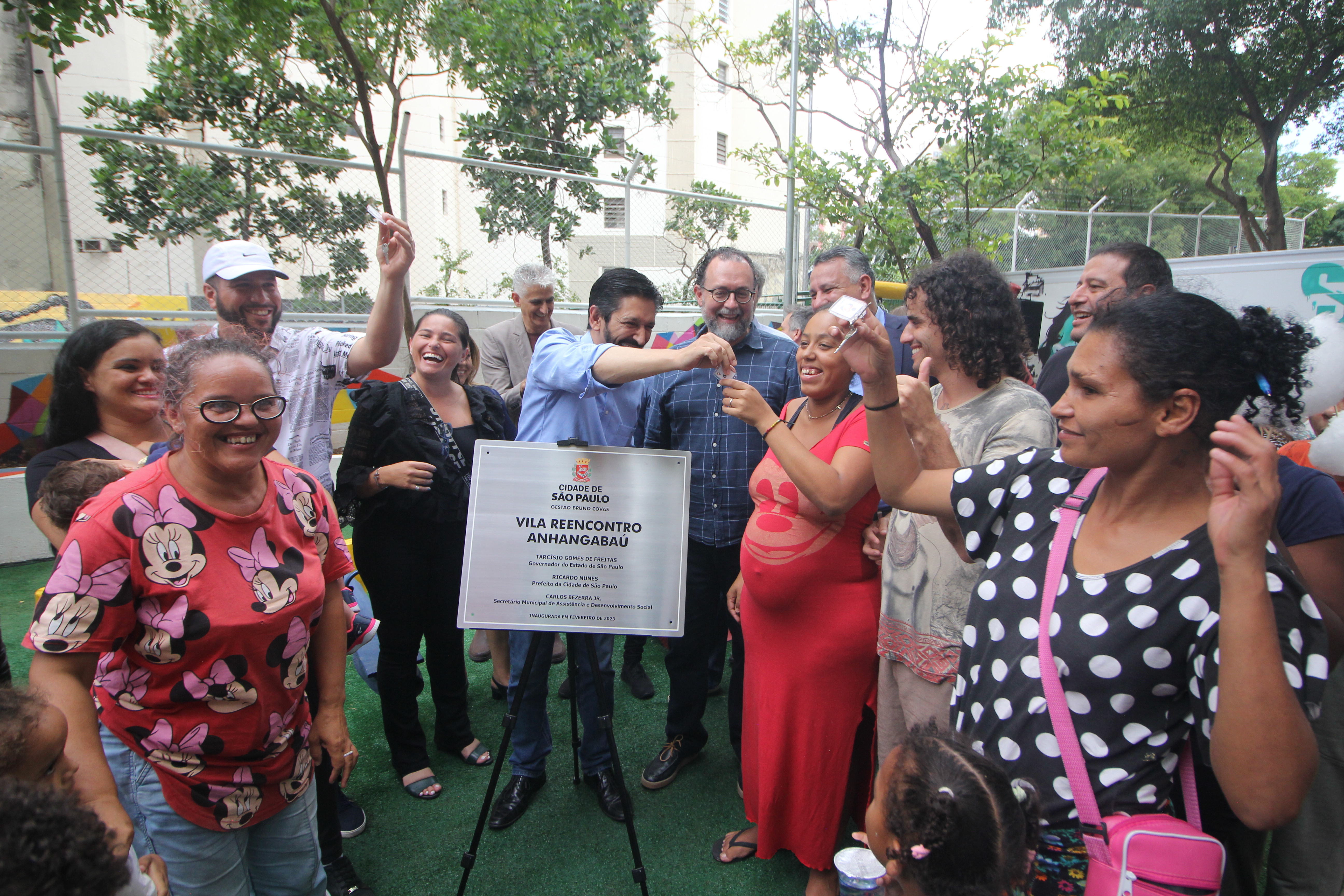 11 pessoas, entre elas o Prefeito Ricardo Nunes e o Secretário Carlos Bezerra Jr., com a placa do novo serviço Vila Reencontro 'Anhangabaú', entregando as chaves das casas às famílias.